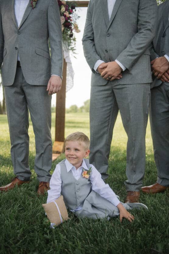 A young boy in a gray suit sits on grass, smiling, holding a ring bearer pillow. Three men in matching gray suits stand behind him, their faces out of frame. The background shows greenery and part of a wooden arch with flowers. by Justin Salem Meyer