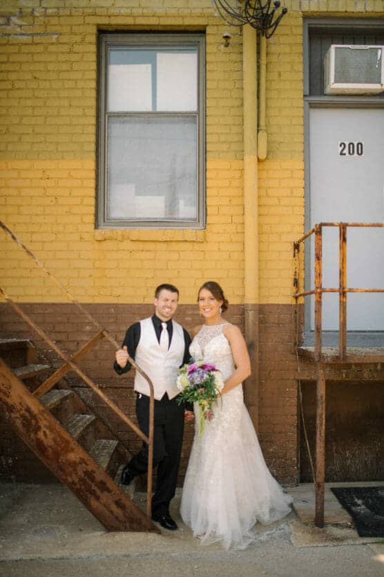 A bride in a white gown holds a bouquet and stands smiling next to a groom in a black suit and white vest. They are by a rusty metal staircase against a yellow and brown brick building with the number 200 visible. by Justin Salem Meyer