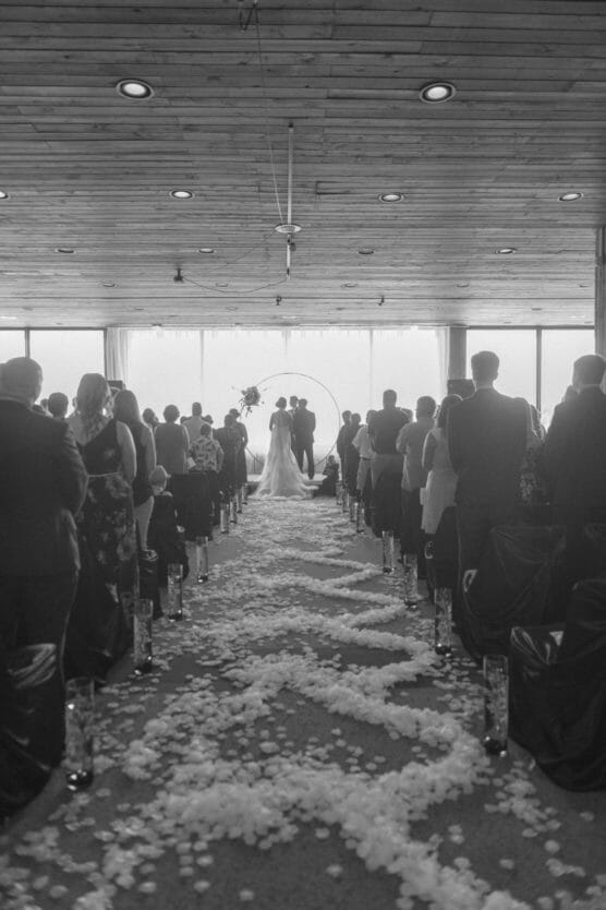 A black and white photo of a wedding ceremony. A couple stands at the altar in front of large windows, surrounded by guests seated on either side of an aisle covered in flower petals. The ceiling is wooden with round lights. by Justin Salem Meyer