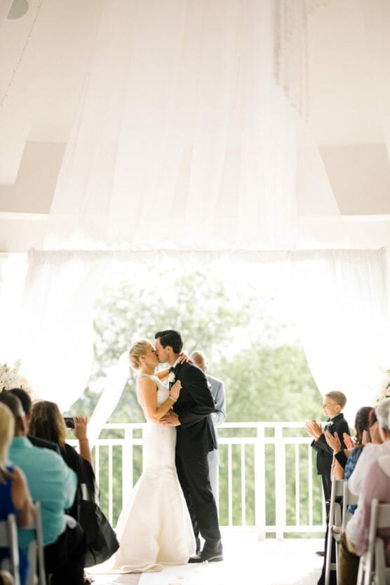A bride and groom share their first kiss at an indoor wedding ceremony, surrounded by seated guests who applaud. The couple stands beneath white drapes with natural light streaming in behind them. by Justin Salem Meyer