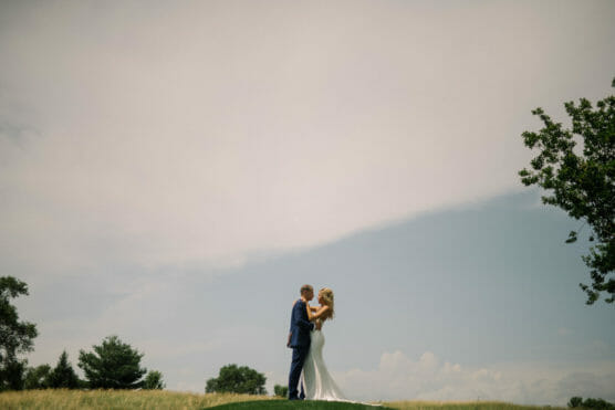 A bride and groom stand close together on a grassy hill under a vast, cloudy sky, surrounded by distant trees, creating a romantic and serene outdoor scene. by Justin Salem Meyer