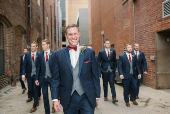 A smiling groom in a navy suit and red bow tie walks in front of his groomsmen, who are also dressed in suits, down a brick alleyway. by Justin Salem Meyer
