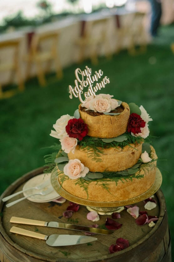 A three-tier savory pie “cake” decorated with pink and red flowers, greenery, and a topper reading Our Adventure Continues, sits on a glass stand atop a wooden barrel, with cake-serving utensils and rose petals nearby. by Justin Salem Meyer