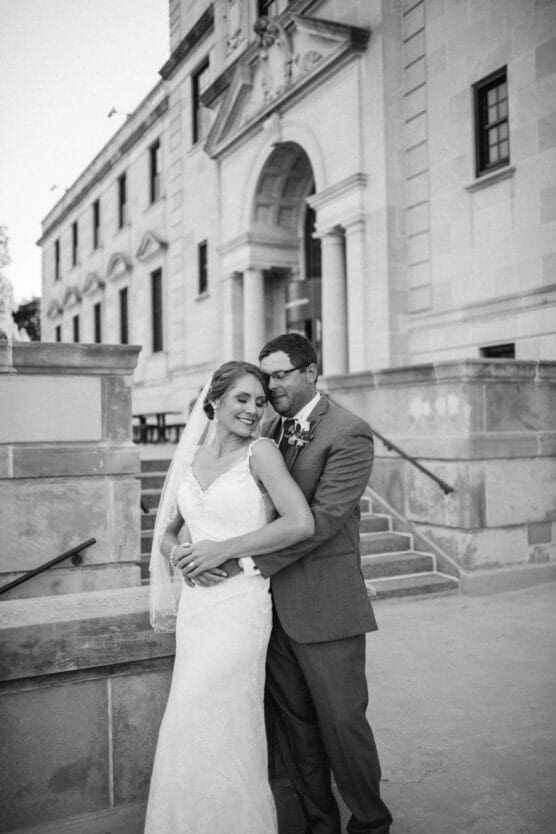 A bride and groom embrace and smile outside a stone building, possibly a courthouse or church. The bride wears a lace gown and veil, and the groom is in a suit. The setting appears formal and celebratory. by Justin Salem Meyer
