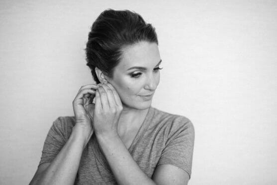 Black and white photo of a woman with styled hair, wearing a short-sleeved shirt, looking down as she puts on an earring with both hands against a plain background. by Justin Salem Meyer