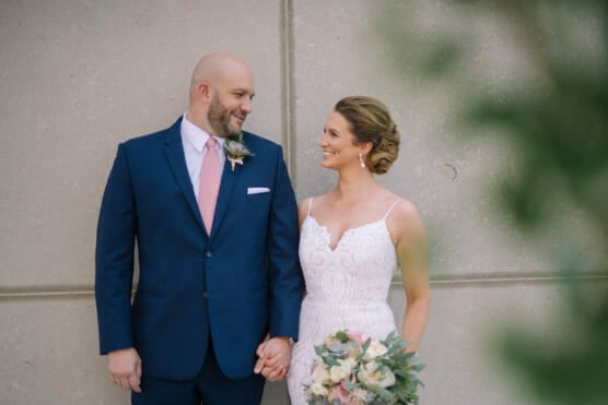 A bride in a white dress and a groom in a navy suit with a pink tie hold hands and smile at each other, standing in front of a light-colored wall, with greenery blurred in the foreground. by Justin Salem Meyer