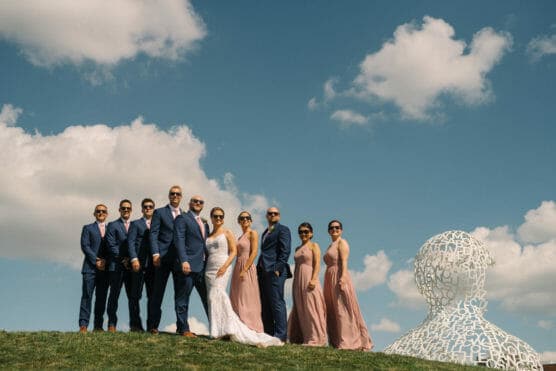 A wedding party poses outdoors on grass under a blue sky with clouds. The group includes men in blue suits and women in blush dresses, all wearing sunglasses. A large white sculpture made of letters is on the right. by Justin Salem Meyer