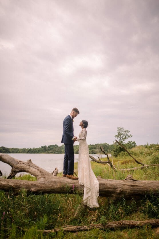 A bride and groom stand facing each other on a large fallen tree beside a lake, holding hands. The groom wears a navy suit; the bride wears a long lace dress with a train. The sky is overcast, and grass surrounds them. by Justin Salem Meyer
