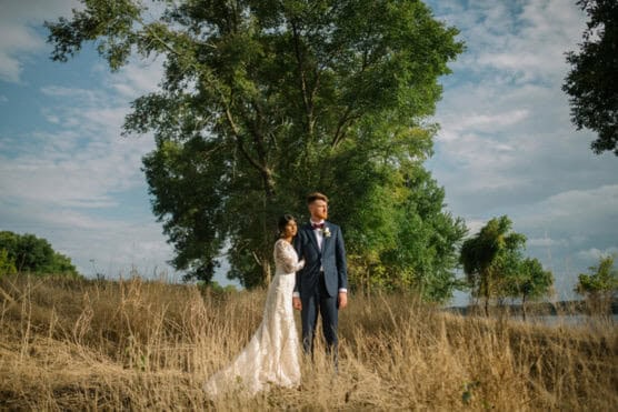 A bride in a white dress and a groom in a navy suit stand together in a sunlit field with tall, dry grass and green trees under a partly cloudy sky. by Justin Salem Meyer