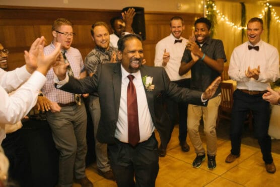 A man in a suit and red tie dances joyfully at a celebration, surrounded by clapping and smiling people in semi-formal attire. The room is warmly lit, with string lights in the background. by Justin Salem Meyer