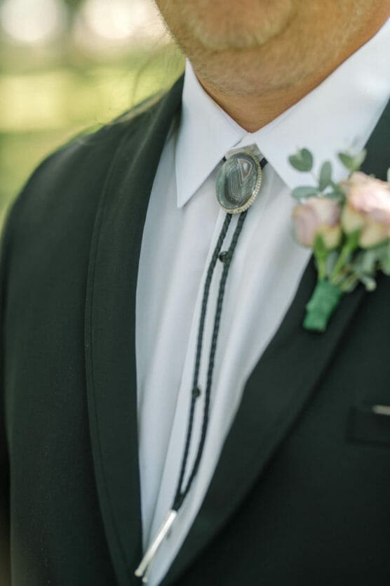 A close-up of a person wearing a dark suit with a white shirt, bolo tie, and a boutonnière of small pink flowers pinned to the lapel. The image is taken outdoors with blurred greenery in the background. by Justin Salem Meyer