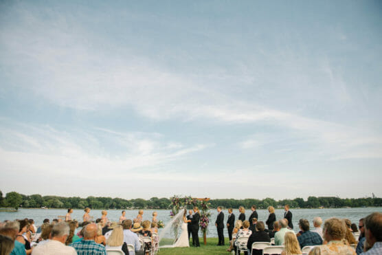 Outdoor wedding ceremony by a lake, with guests seated facing a couple at an altar. Bridesmaids in light dresses and groomsmen in dark suits stand on either side under a clear blue sky. by Justin Salem Meyer