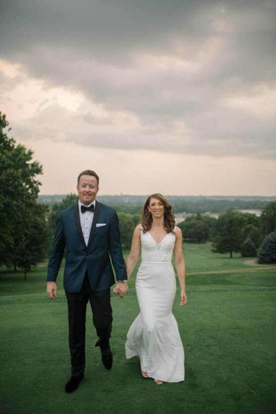 A smiling bride and groom walk hand-in-hand on a green lawn, dressed in a white gown and a dark tuxedo, with trees and a cloudy sky in the background. by Justin Salem Meyer