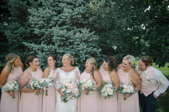 A bride in a white gown stands with six bridesmaids and one groomsman wearing blush pink outfits, all holding bouquets and smiling together in front of a large evergreen tree outdoors. by Justin Salem Meyer