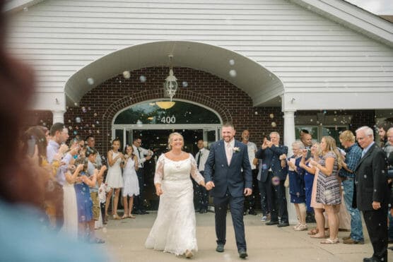 A bride in a white gown and a groom in a navy suit walk hand in hand outside a building as guests surround them, smiling, clapping, and blowing bubbles to celebrate their wedding. by Justin Salem Meyer