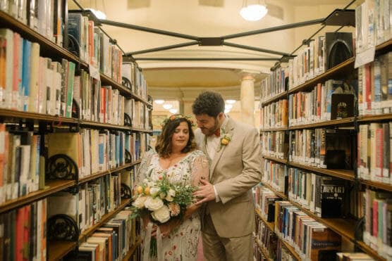 A bride and groom stand closely together between library bookshelves, the bride holding a bouquet and wearing a floral dress with a flower crown, while the groom in a tan suit looks at her affectionately. by Justin Salem Meyer