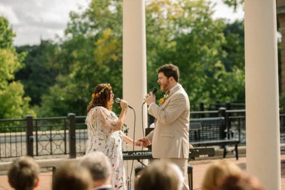 A couple stands holding hands and singing into microphones during an outdoor ceremony, surrounded by greenery and sunlight, with guests seated in the foreground. by Justin Salem Meyer