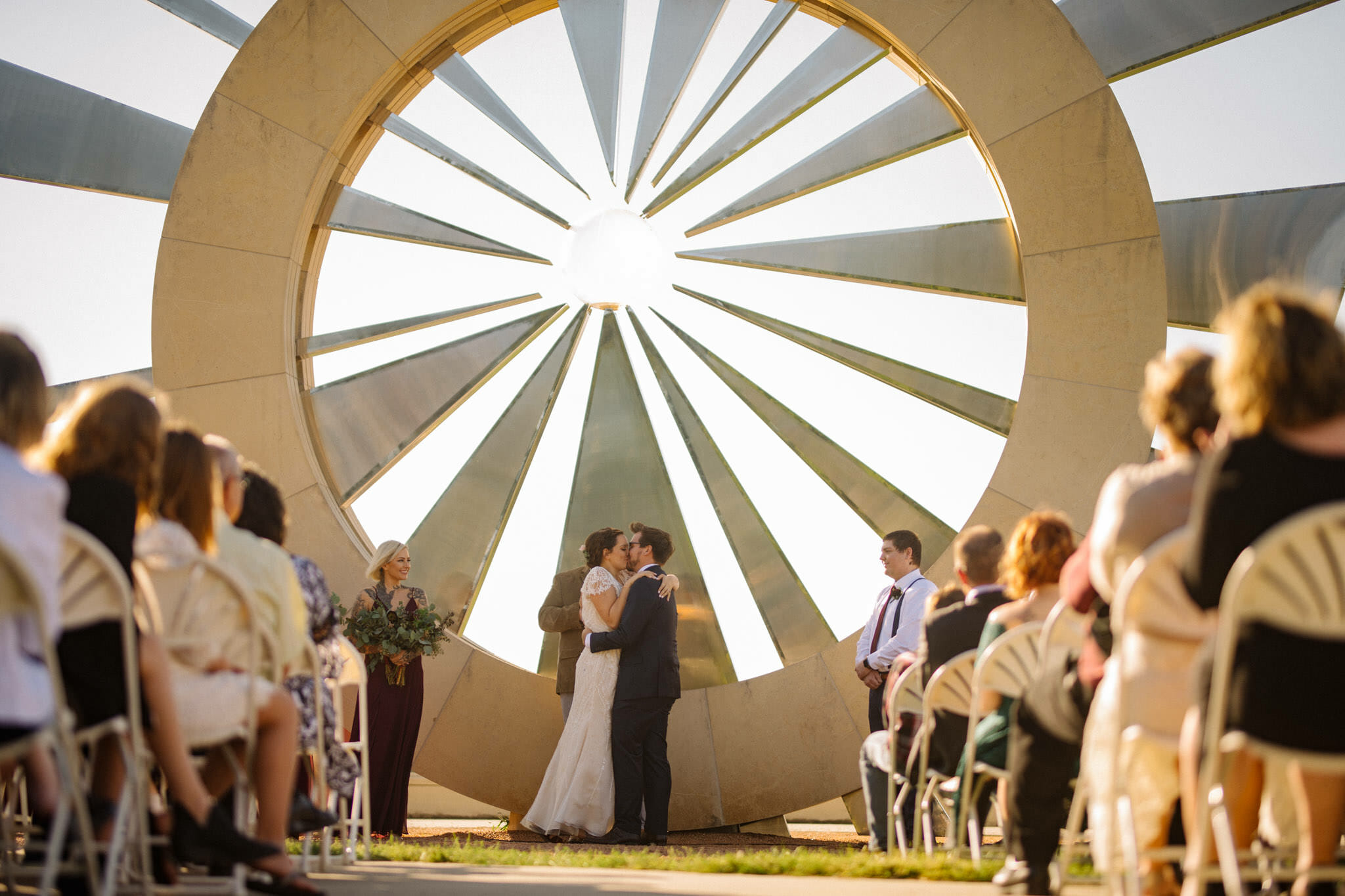 Wedding day couple during their first kiss below Shattering Silence sculpture overlooking downtown Des Moines, Iowa