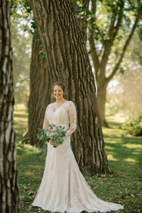 A bride in a long-sleeved lace wedding dress stands outdoors, holding a bouquet of flowers, smiling, with tall trees and sunlight filtering through green leaves in the background. by Justin Salem Meyer