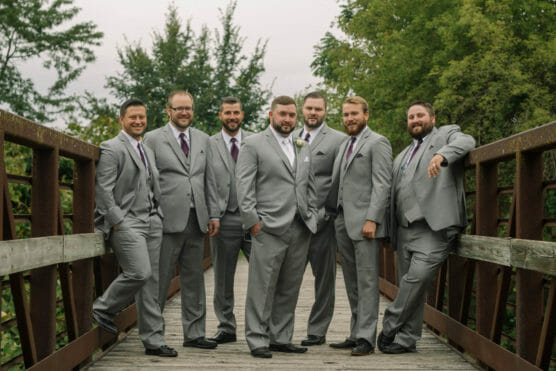 Seven men wearing light gray suits and purple ties stand together on a wooden bridge outdoors, posing for a group photo with trees in the background. by Justin Salem Meyer