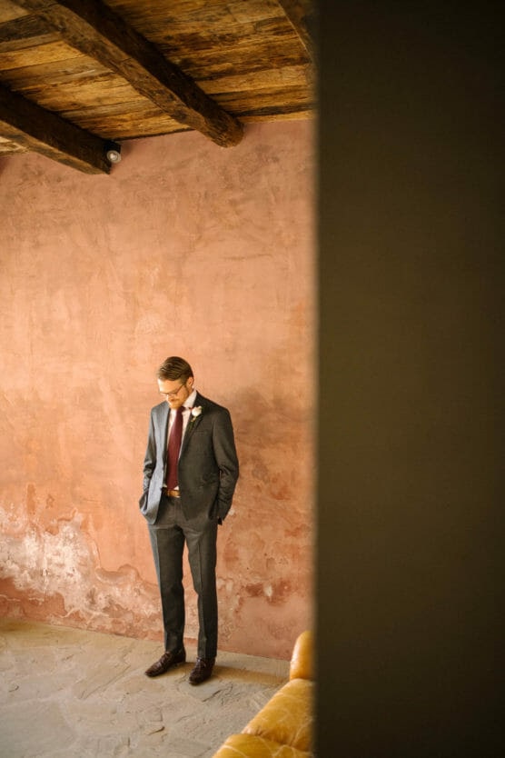 A man in a gray suit with a red tie and boutonnière stands against a textured, peach-colored wall, looking down with his hands in his pockets. Wooden beams are visible on the ceiling above him. by Justin Salem Meyer
