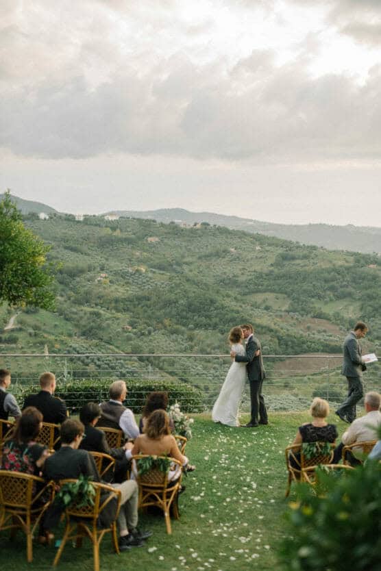 A bride and groom share a kiss during an outdoor wedding ceremony on a grassy hilltop, surrounded by seated guests and overlooking a scenic, green valley with rolling hills under a cloudy sky. by Justin Salem Meyer