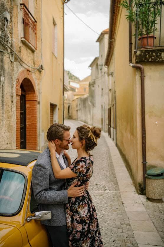 A couple embraces lovingly next to a yellow vintage car on a narrow cobblestone street lined with warm-colored buildings in a quaint European town. by Justin Salem Meyer