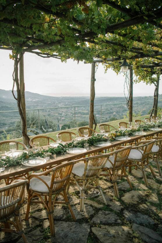 A long wooden table with wicker chairs is set for an outdoor meal under a leafy pergola, overlooking a scenic hillside landscape with mountains in the distance. by Justin Salem Meyer