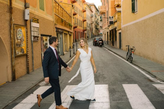 A bride in a white dress and a groom in a dark suit hold hands and smile as they walk across a crosswalk on a colorful, narrow street lined with buildings and a bicycle. by Justin Salem Meyer