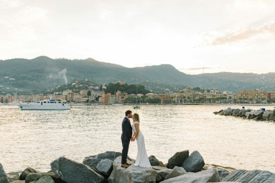 A couple dressed in wedding attire stands holding hands on large rocks by the water, with a scenic coastal town, mountains, and a boat in the background at sunset. by Justin Salem Meyer