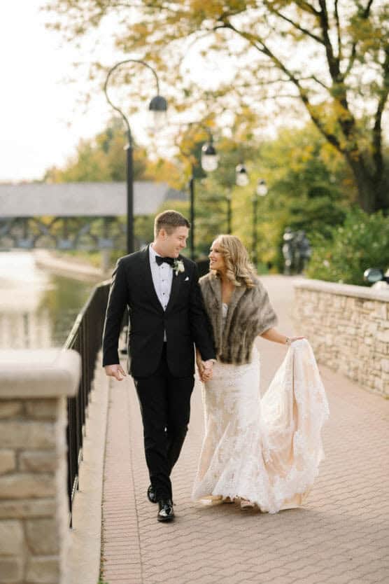 A bride in a lace gown and fur shawl walks hand in hand with a groom in a black tuxedo along a stone walkway by a river, with trees and a bridge in the background. by Justin Salem Meyer