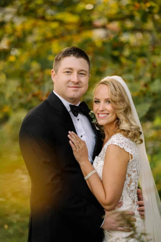 A bride in a white lace dress and veil stands smiling next to a groom in a black tuxedo, outdoors with greenery in the background. They are embracing and looking at the camera. by Justin Salem Meyer