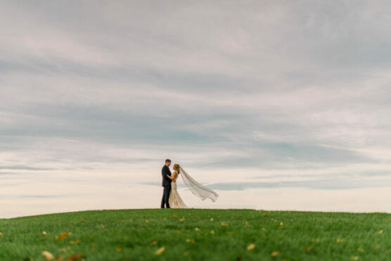 A bride and groom stand together on a grassy hill under a cloudy sky. The bride’s veil flows behind her in the wind, and they are holding each other, creating a romantic and serene scene. by Justin Salem Meyer