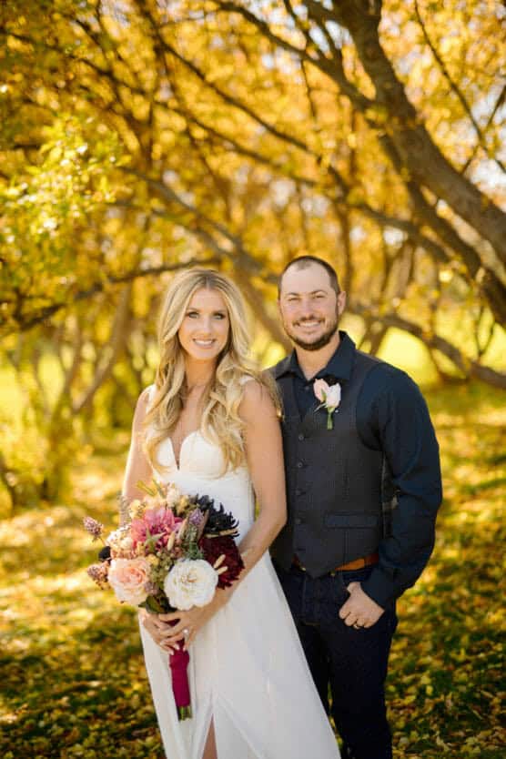 A smiling bride in a white dress holds a bouquet of flowers while standing next to a smiling groom in a dark shirt and vest. They are outdoors under golden autumn trees with sunlight filtering through. by Justin Salem Meyer