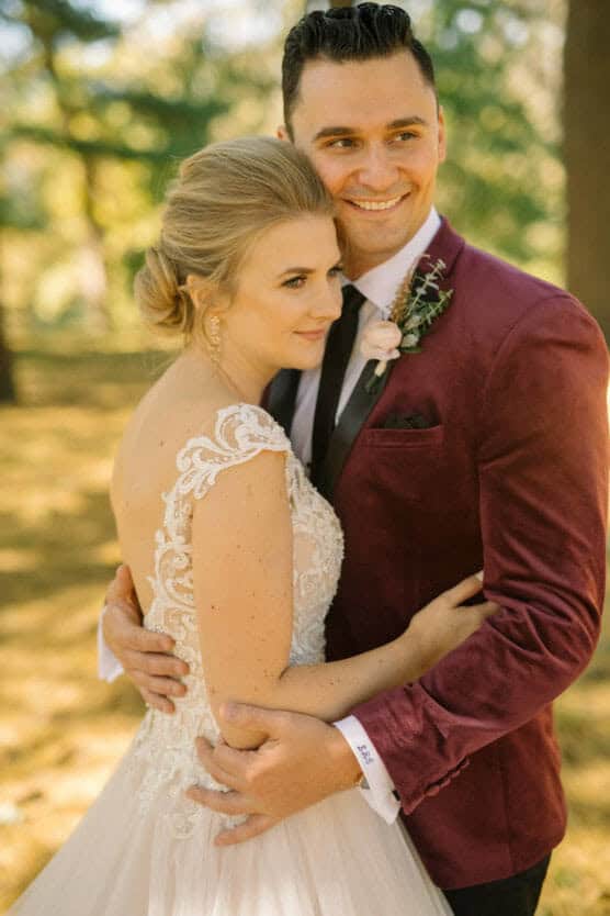 A bride in a lacy white wedding dress and a groom in a burgundy jacket embrace outdoors, smiling happily, with sunlight filtering through trees in the background. by Justin Salem Meyer