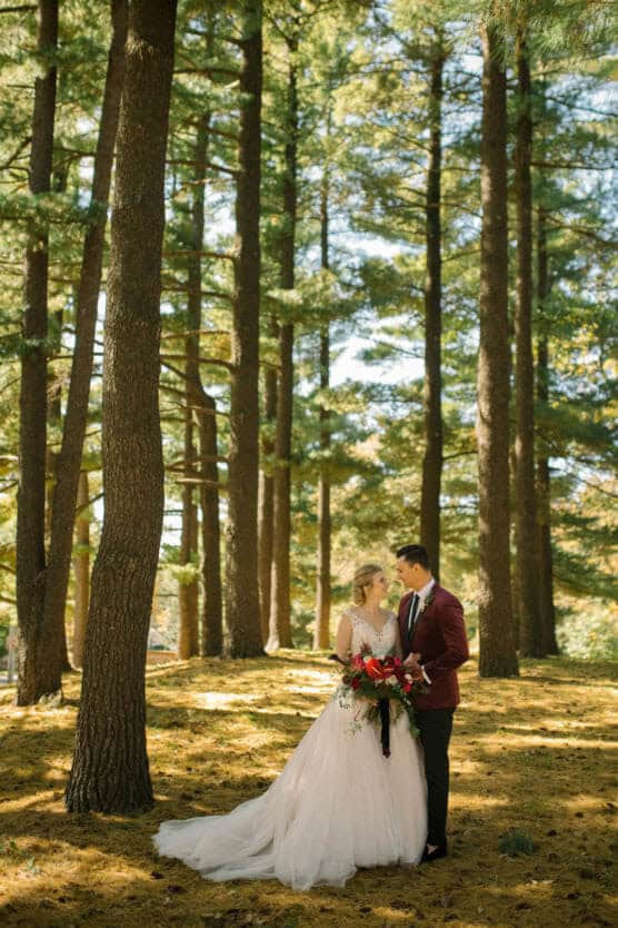 A bride and groom stand together in a sunlit forest, surrounded by tall pine trees. The bride wears a white gown and holds a bouquet of flowers, while the groom wears a dark suit with a burgundy jacket. by Justin Salem Meyer