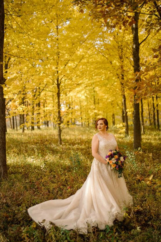 A woman in a white wedding gown stands in a sunlit forest with vibrant yellow autumn leaves, holding a colorful bouquet and gazing to the side. by Justin Salem Meyer
