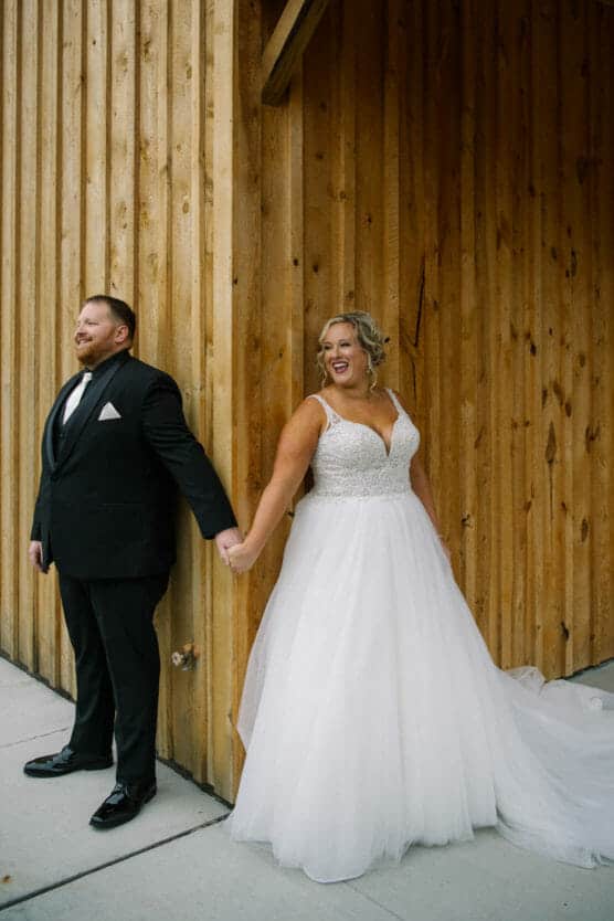 A bride in a white gown and a groom in a black tuxedo stand back to back, holding hands and smiling, next to a wooden wall on their wedding day. by Justin Salem Meyer