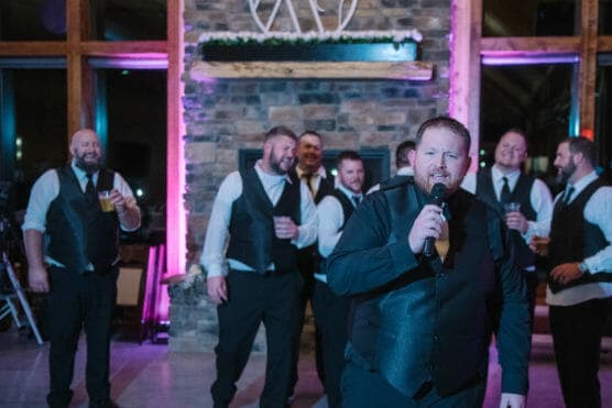 A man in a black vest and tie sings into a microphone, while a group of men in matching formal attire stand behind him, smiling and holding drinks, in front of a stone wall with purple lighting. by Justin Salem Meyer