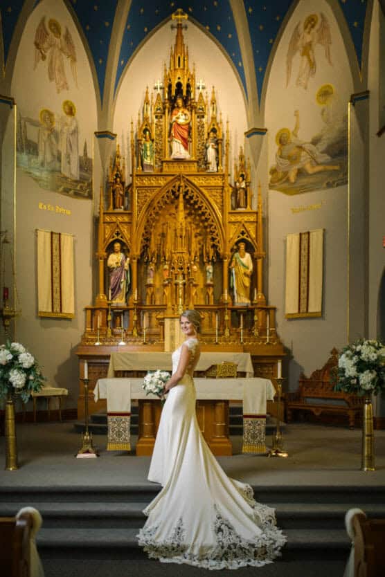 A bride in a white gown with a long lace train stands facing sideways, holding a bouquet in a church with ornate golden altar, statues, and arched blue ceiling. by Justin Salem Meyer