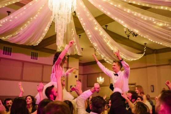 A bride and groom are lifted on chairs by guests at a lively wedding reception, surrounded by cheering friends and family under draped fabric and string lights with a grand chandelier overhead. by Justin Salem Meyer