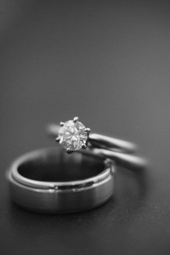 A close-up black and white photo of two rings: a diamond engagement ring rests on top of a plain wedding band, both on a smooth, dark surface. by Justin Salem Meyer