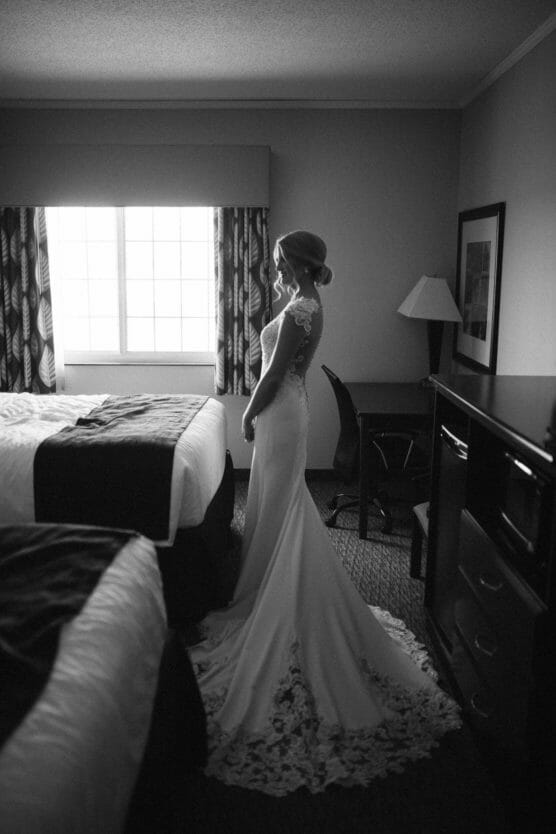 A bride in a long, lace-trimmed gown stands in a softly lit hotel room, facing a window with sheer curtains, as light gently highlights her figure. The room contains two beds, a desk, and a lamp. by Justin Salem Meyer