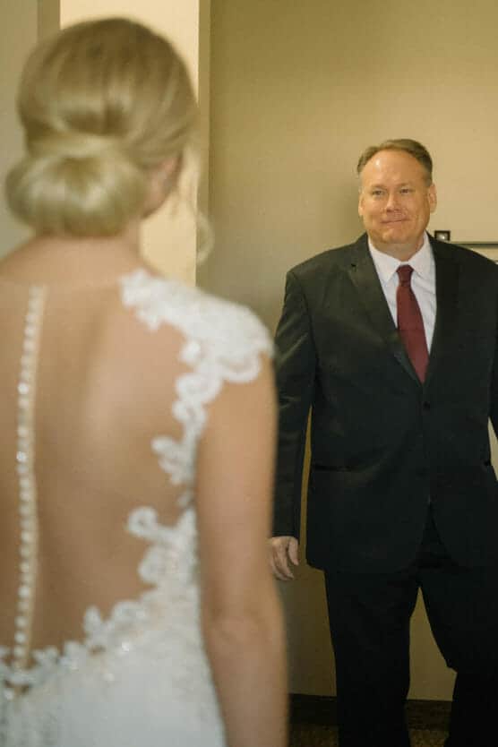 A woman in a white lace wedding dress faces a smiling man in a dark suit and red tie, who looks pleasantly surprised. The woman’s back is to the camera, showcasing her dress’s sheer and buttoned back. by Justin Salem Meyer