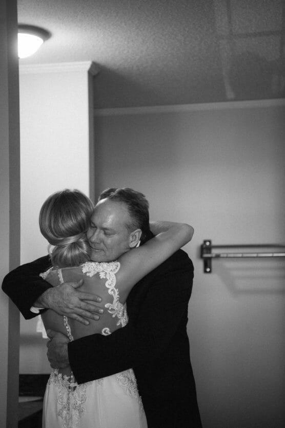 A black and white photo of a bride in a lace wedding dress hugging an older man in a suit. They embrace warmly in a room with plain walls and a shelf in the background. by Justin Salem Meyer