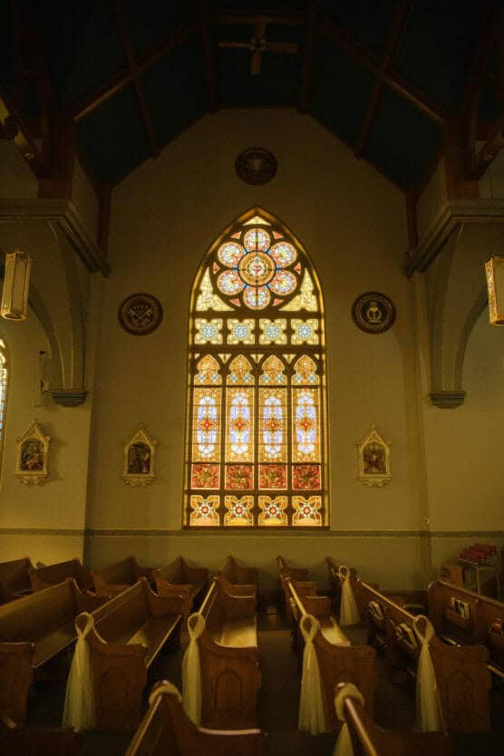 A church interior with wooden pews decorated with white ribbons, facing a large ornate stained glass window that glows with warm light. The walls are adorned with religious artwork and high, vaulted ceilings. by Justin Salem Meyer