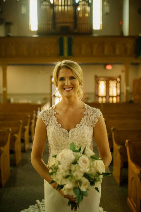 A bride in an elegant lace wedding dress holds a bouquet of white flowers, smiling as she stands in the aisle of a warmly lit church with wooden pews and stained glass in the background. by Justin Salem Meyer