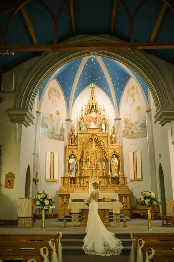 A bride in a white gown stands alone facing an ornate, gold altar in a church with high arched ceilings, blue accents, religious artwork, and floral arrangements on either side. by Justin Salem Meyer