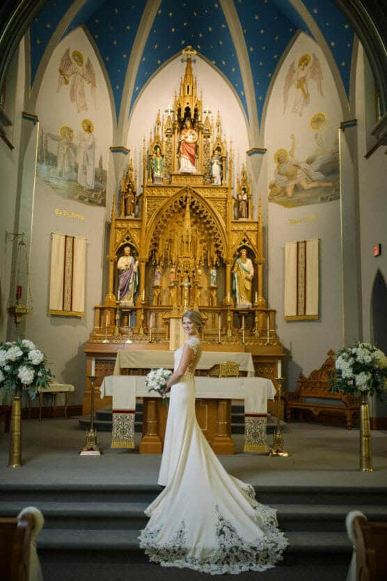 A bride in a white gown stands smiling in front of an ornate altar inside a church with blue arched ceilings, gold details, and religious statues. Large floral arrangements are on either side of her. by Justin Salem Meyer
