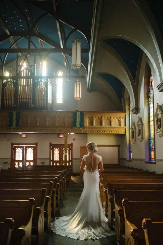 A bride in a white gown with a long train stands in the aisle of a sunlit church, facing the altar. Stained glass windows and wooden pews surround her under a high, arched ceiling. by Justin Salem Meyer